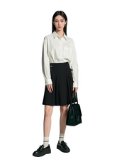 Woman wearing a white shirt and black pleated skirt with a green handbag on a gray background