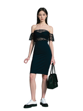 Woman wearing a dark green off-shoulder dress with lace details, holding a matching handbag, standing on a gray background.